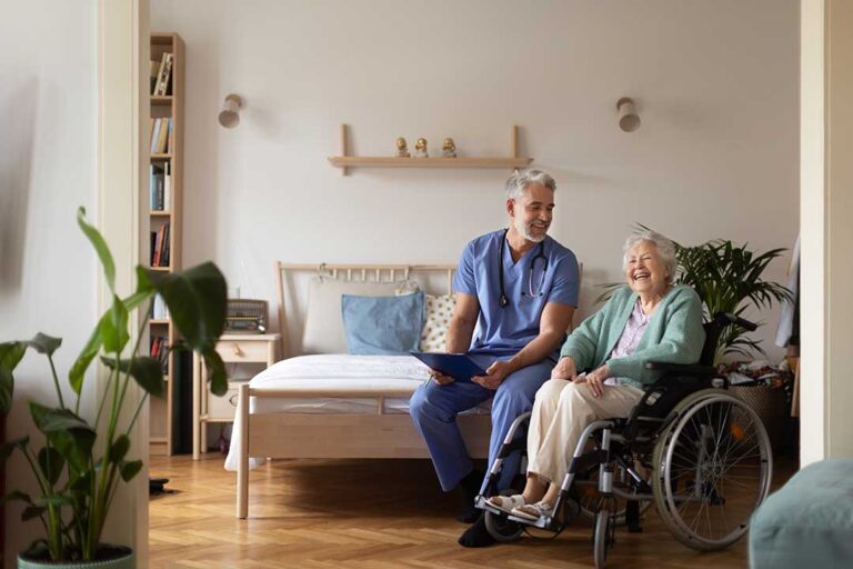 An older adult chatting with neighbors in a bright assisted living common room, surrounded by comfortable furniture and plants.